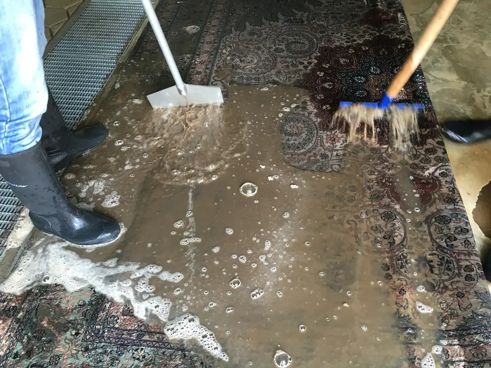 Braunes Waschwasser läuft aus einem hellen Orientteppich, zwei Mitarbeiter schieben es mit Wasserschiebern ab